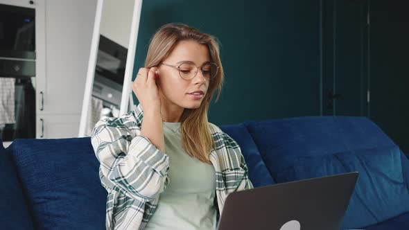 Young Blonde Woman Putting on Wireless Earphones and Working on Laptop Sitting Cozily on Sofa Ta alt