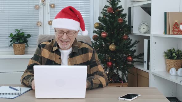 A Middleaged Man in a Santa Claus Hat Happily Communicates Via Video Communication on a Laptop and