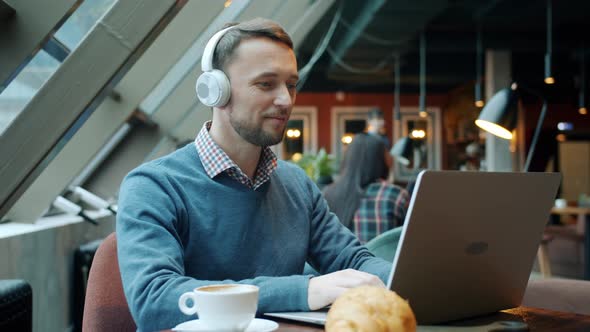 Guy Wearing Headphones Listening To Music and Working with Laptop in Cafe alt