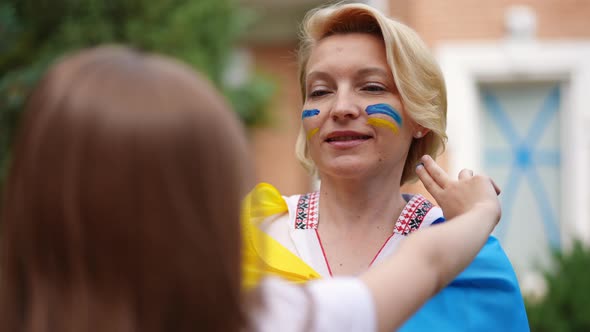 Portrait of Proud Mother Smiling As Daughter Drawing Ukrainian Flag Colors on Cheeks Leaving alt