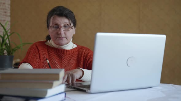 Modern Elderly Woman Writes Information in a Notebook While Studying Online Using Modern Technology alt