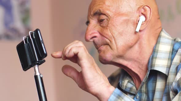 Caucasian pensioner 70-75 years old in headphones at home holds a tripod with a smartphone and click alt