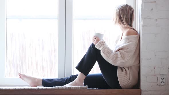 young girl in sweater and jeans is sitting at window with cup of coffee at home alt