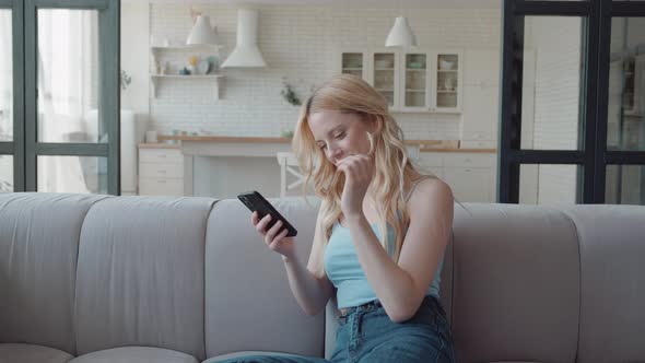 Happy Blonde Woman Using Smartphone Sitting on the Couch in Cozy Stylish Living Room Chatting and alt