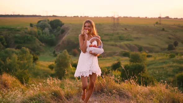 Beautiful sexy blonde girl in white dress posing in a field at sunset with a basket of fruit alt