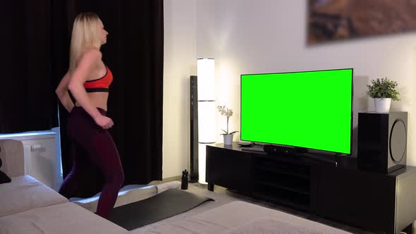 A young beautiful woman does a fitness dance in front of a TV with a green screen in an apartment alt