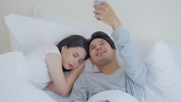 Asian attractive funny male selfies take picture of sleeping girlfriend on bed in bedroom at home. alt