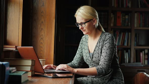 Business Woman in Glasses Works with a Laptop in a Library or a Luxurious Office. In the Background