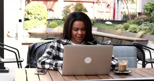 Happy stylish African American female student using laptop computer, drinking coffee.
