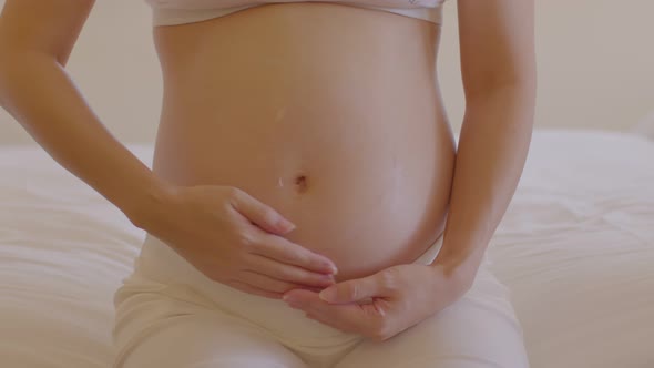 Happy Pregnant Woman sitting on bed holding and stroking apply cream on big belly alt