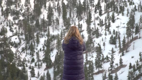 Blonde Girl Raises Her Hands Up Against the Background of Snowy Pine Forest in the Mountain alt