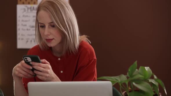 Blond hair woman in red shirt working at home office on laptop computer and speaking by mobile phone alt