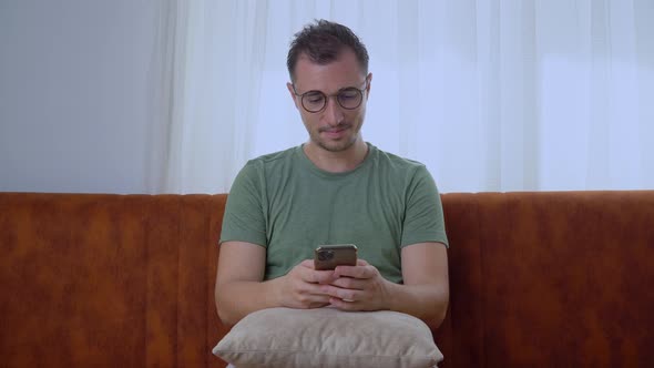 A Man with Glasses with a Smartphone Sits at Home on the Couch and Corresponds in the Phone