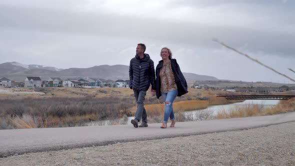 Young couple holding hands as they walk along a paved river trail on a windy winter afternoon in a r alt