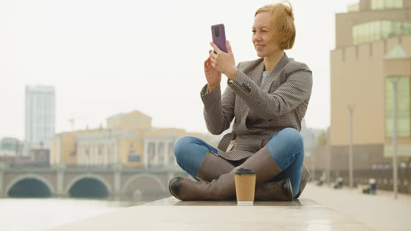 40Yearold Beautiful Woman Sits on the Embankment and Takes a Photo of the Cityscape with Her alt