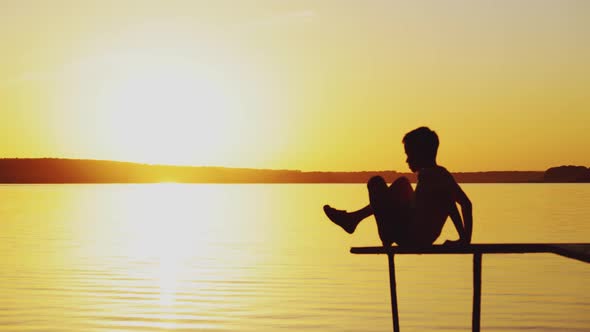 Little Boy is Sitting in a Lotus Position on the Edge of a Bridge Above a River alt