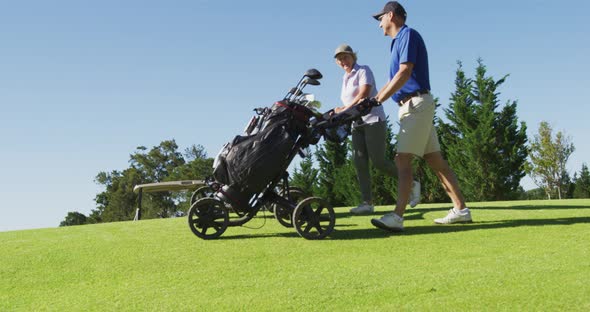 Caucasian senior couple walking with their golf bags at golf course on a bright sunny day alt