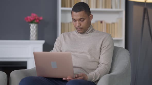 Middle Shot Portrait of Concentrated Young African American Man Sitting on Armchair Typing on Laptop alt