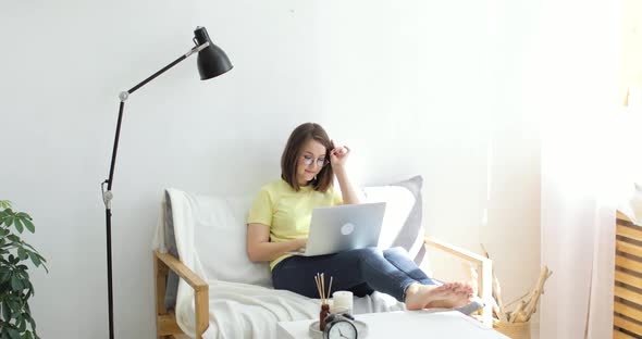 Young Beautiful Woman with Laptop is Sitting in a Large White Armchair Next to a Lamp and Chatting