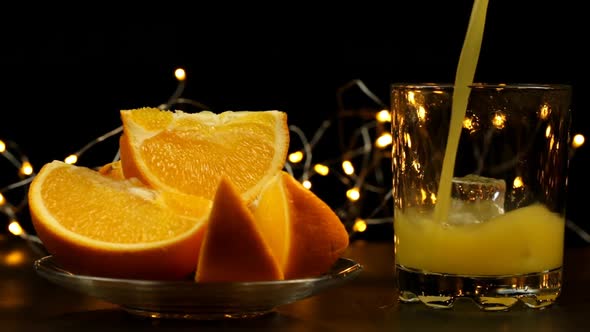 Juice Is Poured Into A Glass With Ice, Several Sliced Oranges In A Plate On A Wooden Table