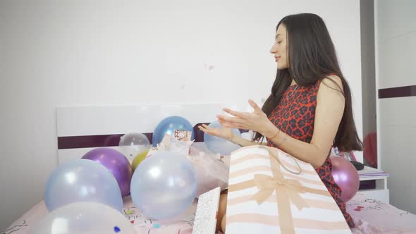 Side View of Woman Sitting on Bed with Colorful Balloons and Opens Confetti Bomb Birthday Card alt