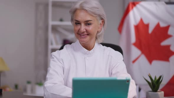Joyful Mature Smiling Woman Talking Looking at Camera with Canadian Flag at Background alt