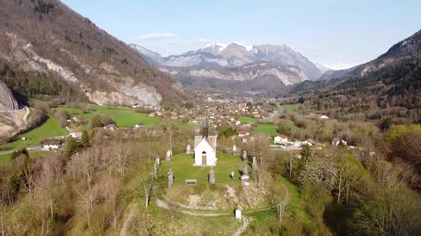 Drone Shot (facing forwards,ing up and back) of a Chapel on top of a Hill in front of Alpine Mountai alt