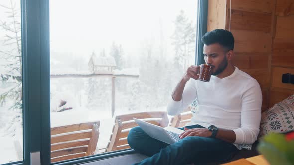 Young Indian Man Drinking Coffee and Working on Laptop on Winter Day alt