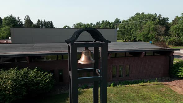 Single gold bell in a sturdy structure outside a brick building on a sunny day with shadows on grass