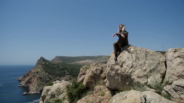 A Lonely Hippie Traveller with Long Blond Hair Sits on a Rock in the Mountains alt