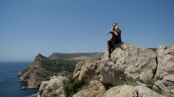 A Lonely Hippie Traveller with Long Blond Hair Sits on a Rock in the Mountains