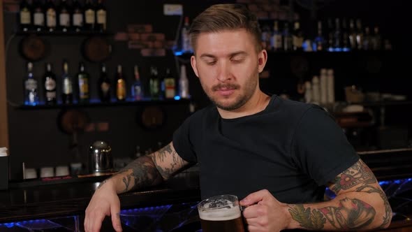 A Bearded Young Man with Tattoos Drinking Beer at Bar or Pub alt