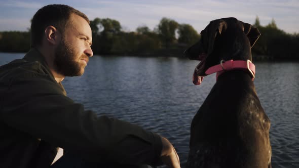 Young Guy Sitting with Dog on the Pier and Admiring the Beauty of the Lake alt