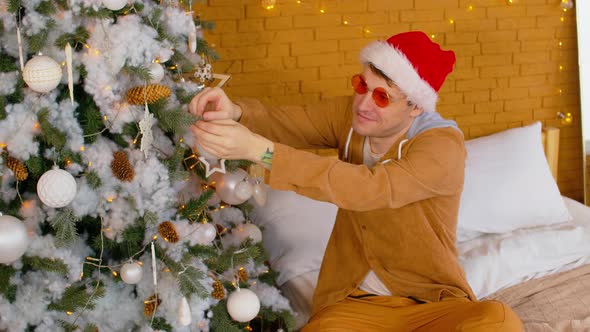 Man in Santa Hat and Fashionable Glasses Sitting on Bed and Decorating Christmas Tree alt