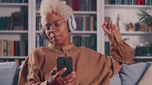 African Woman Relaxing on Sofa Holds Smartphone Listens Song Through Headphones alt
