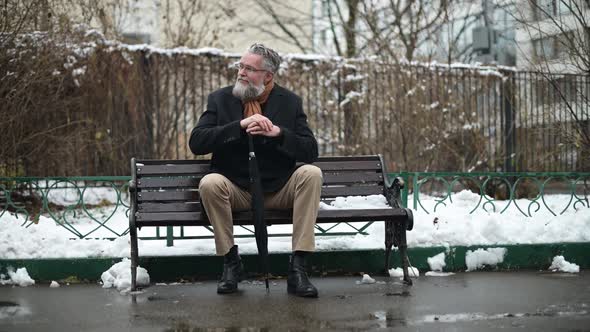 An adult gray-haired respectable man with glasses sits on a park bench alt