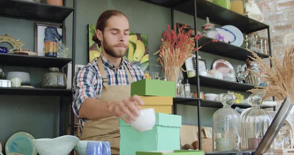 Bearded Man in Casual Clothes and Apron Sitting at Workplace in Beautiful Gift Shop alt