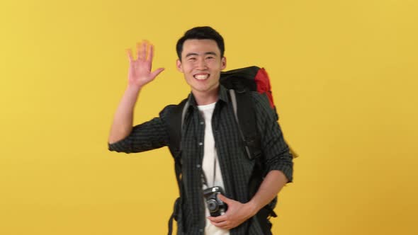 Happy young Asian tourist man with backpack clicks photo on camera and waves his hand
