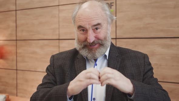 Smiling old bearded man showing hands sign heart shape looking at camera. alt