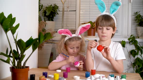 a Little Girl and a Boy with the Ears of Easter Hares Paint Eggs for the Holiday with Brushes alt