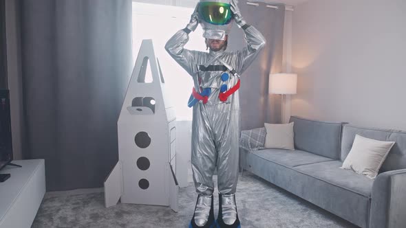 Middleaged Man in an Astronaut Suit Stands in Living Room Next to a Cardboard Model of a Space alt