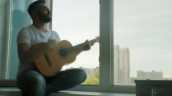 Handsome Young Man Playing Guitar and Singing Having Fun Sitting on Window Sill at Home alt