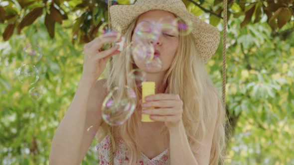 Smiling blonde woman wearing straw hat, makes soap bubbles sitting on the swing in the home garden