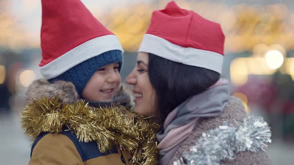 Closeup Happy Middle Eastern Mother and Son in Christmas Hats Rubbing Noses Turning Looking at alt
