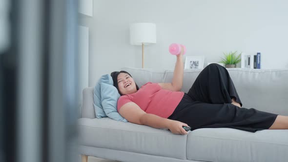 Asian funny oversize big girl try to exercise for health using dumbbell workout while lying on sofa. alt