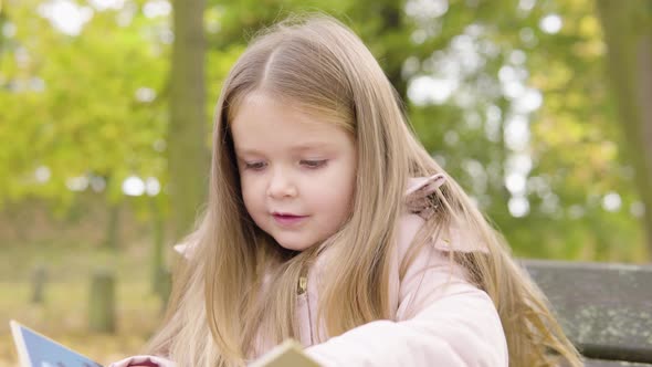 Cute Little Caucasian Girl Reads a Picture Book Out Loud with a Smile As She Sits on a Bench in Park alt