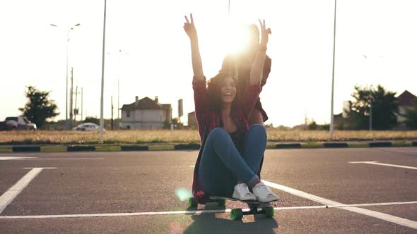 Cheerful Woman Sitting on a Longboard and Raising Her Hands Up Happily While Her Friend is Pushing alt