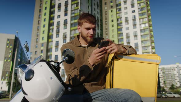 Courier After Delivery Sits on a Scooter and Looks at the Phone in the City alt