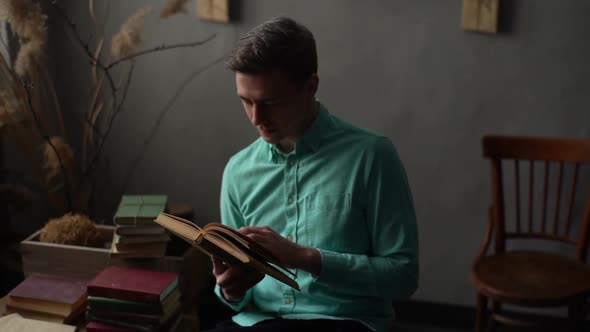 Medium Shot of Focused Young Man Flips Through Pages of Old Frayed Book Sitting in Chair in Dark alt