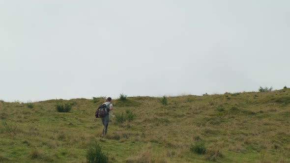 Beautiful Girl with a Backpack Walks Through the Grass on a Green Hill in the Afternoon